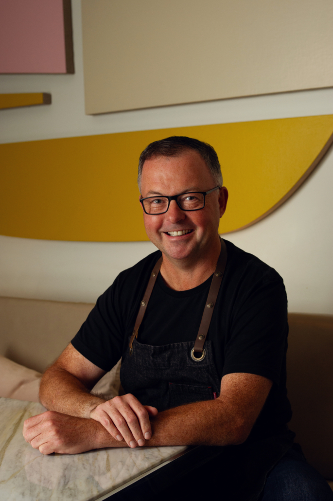 Chef David Hawksworth sits at a table and smiles at the camera.
