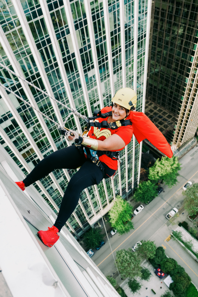 One Drop Zone rappeller hanging off the side of the building partway down in rappelling. She is wearing a red cape, red shirt and red shoes. 